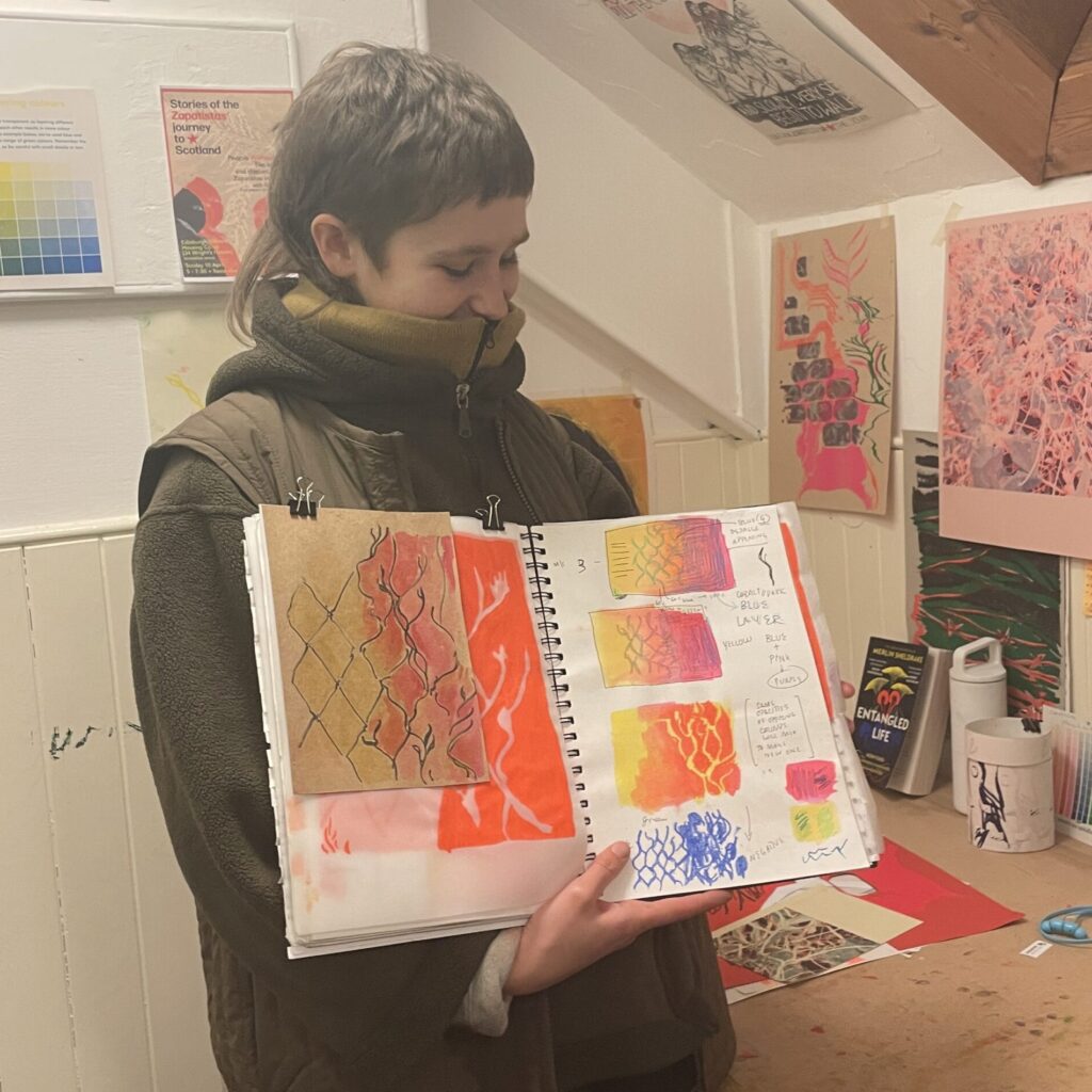 Artist Celeste is holding up a sketchbook full of colourful sketches of chain-link fences. 